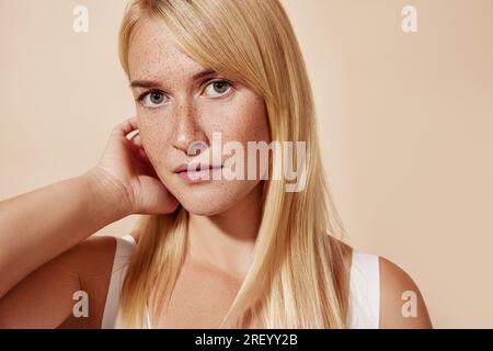 Jeune belle femme blonde avec des taches de rousseur regardant la caméra et ajustant ses cheveux Banque D'Images