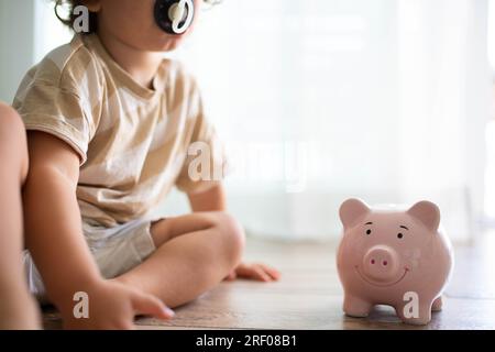 Enfant caucasien avec pacificateur, assis sur le sol près d'une tirelire souriante. Économiser de l'argent pour les enfants concept. Banque D'Images