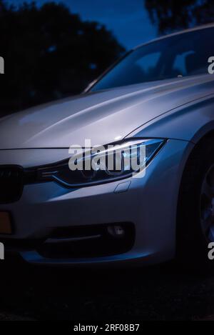 Detailshot d'un phare BMW série 3 de 2012. Pris la nuit, un lampadaire met en valeur le capot et le phare de la voiture argentée. Banque D'Images