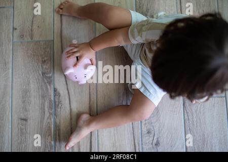 Enfant caucasien avec pacificateur, assis sur le sol près d'une tirelire souriante. Économiser de l'argent pour les enfants concept. Banque D'Images