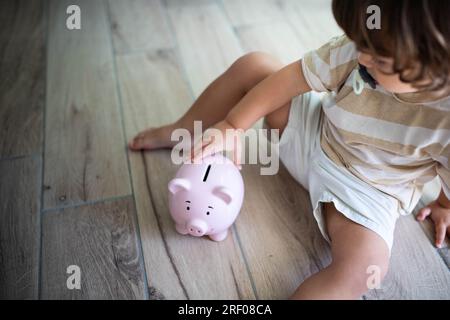 Enfant caucasien avec pacificateur, assis sur le sol près d'une tirelire souriante. Économiser de l'argent pour les enfants concept. Banque D'Images