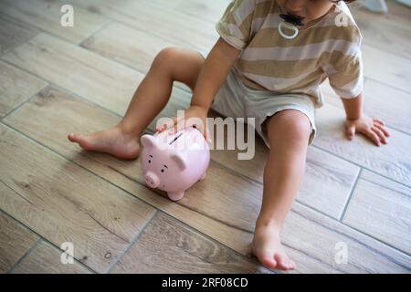 Enfant caucasien avec pacificateur, assis sur le sol près d'une tirelire souriante. Économiser de l'argent pour les enfants concept. Banque D'Images