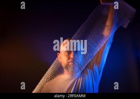 Homme barbu chauve étirant un film à bulles devant son visage. Isolé sur fond rouge foncé. Banque D'Images