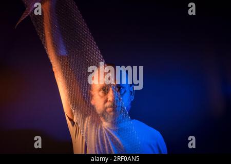 Homme barbu chauve étirant un film à bulles devant son visage. Isolé sur fond rouge foncé. Banque D'Images