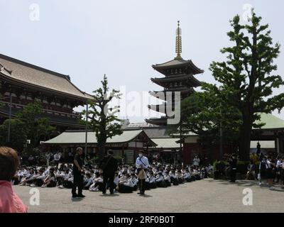 Un grand groupe d'écoliers japonais en uniforme sont assis sur le sol avec une pagode au temple Senso-ji en arrière-plan ; visite touristique à Tokyo. Banque D'Images