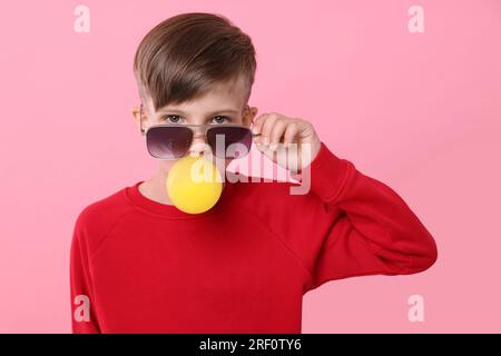 Garçon dans les lunettes de soleil soufflant du bubble gum sur fond rose Banque D'Images