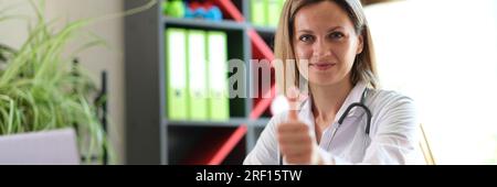 Une femme médecin assise à son bureau dans une clinique médicale et montre le pouce vers le haut. Banque D'Images