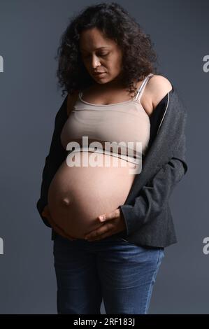 Belle femme dans un blazer et un Jean, touche son ventre enceinte dans le neuvième mois de sa grossesse heureuse sans soucis, sentant des coups de pied de bébé, appréciant tapis Banque D'Images