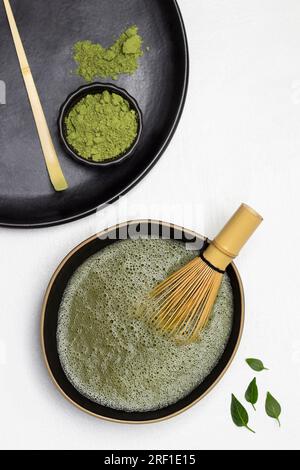 Fouetter en bambou dans un bol avec du thé matcha. Cuillère à mesurer et poudre de thé matcha sur une assiette noire. Pose à plat. Fond blanc Banque D'Images