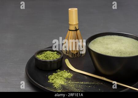 Cuillère doseuse et poudre de thé matcha, thé vert matcha dans un bol et fouet en bambou sur une assiette noire. Espace de copie. Fond gris. Banque D'Images