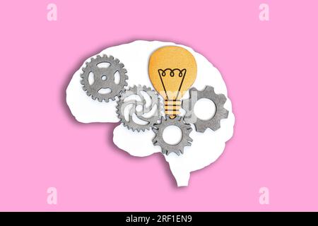 Engrenages en bois et une ampoule placée à l'intérieur d'une forme de cerveau blanc plat isolé sur fond rose. Banque D'Images