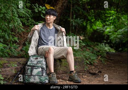 Un randonneur asiatique masculin fatigué et en sueur est assis sur une pierre, se reposant et prenant une pause lors de son voyage de randonnée dans la forêt seul. Banque D'Images