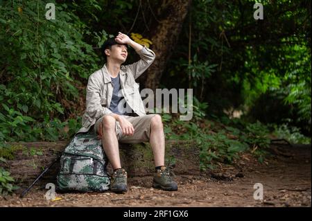 Un randonneur asiatique masculin épuisé et en sueur est assis sur une pierre, se reposant et prenant une pause lors de son voyage de randonnée dans la forêt seul. Banque D'Images