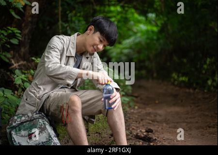 Un randonneur asiatique actif et détendu est assis sur un rocher, buvant de l'eau, tout en se reposant pendant la randonnée dans la forêt. Banque D'Images