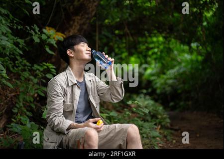 Un randonneur asiatique heureux et détendu est assis sur un rocher, buvant de l'eau, tout en se reposant pendant la randonnée dans la forêt. Banque D'Images