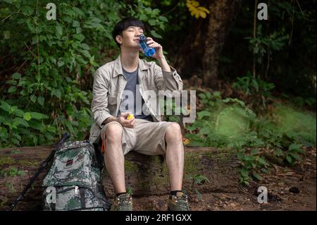 Un randonneur asiatique heureux et détendu est assis sur un rocher, buvant de l'eau d'une bouteille, tout en se reposant pendant la randonnée dans la forêt. Banque D'Images