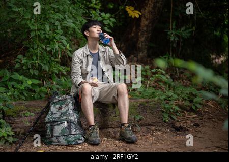 Un randonneur asiatique heureux et détendu est assis sur un rocher, buvant de l'eau d'une bouteille, tout en se reposant pendant la randonnée dans la forêt. pause, repos, détente Banque D'Images