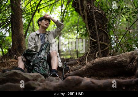 Un randonneur asiatique masculin épuisé et en sueur est assis et prend une pause tout en randonnant dans la forêt verdoyante. Banque D'Images