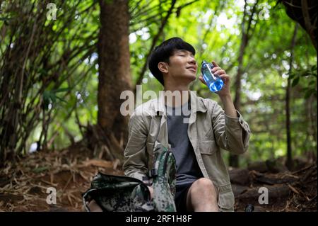 Un randonneur asiatique masculin détendu et heureux est assis, buvant de l'eau et prenant une petite pause pendant son voyage de randonnée dans la forêt verdoyante. Banque D'Images