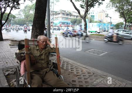 Vieil homme vietnamien dormant dans un fauteuil roulant sur le côté bruyant de la route dans le District 1, Ho Chi Minh ville, Vietnam, Asie du Sud-est Banque D'Images