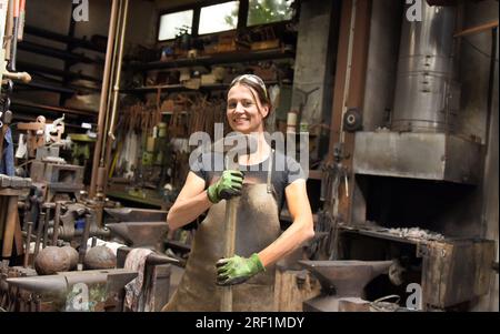 27 juillet 2023, Saxe, Hohenprießnitz : Marika Widdermann, 39 ans, forgeron et designer de métal, se tient en riant avec un marteau dans la forge de son père Roger Widdermann. Avec leur forge, l’équipe père-fille est l’une des rares entreprises artisanales pratiquant encore cet artisanat ancien en Allemagne. En plus des objets utilitaires individuels et des bijoux, ils créent également des meubles forgés, des bols à feu et des boules de feu pour le recyclage des résidus de cire, qui sont particulièrement populaires pendant les mois d'été, pour les clients au pays et à l'étranger. Photo : Waltraud Grubitzsch/dpa Banque D'Images