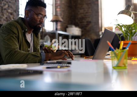 Homme d'affaires occasionnel afro-américain utilisant un ordinateur portable et mangeant de la salade à emporter assis au bureau dans le bureau Banque D'Images