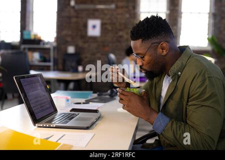 Homme d'affaires occasionnel afro-américain utilisant un ordinateur portable et mangeant de la salade à emporter assis au bureau dans le bureau Banque D'Images