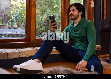 Homme indien assis par la fenêtre et utilisant smartphone à la maison Banque D'Images
