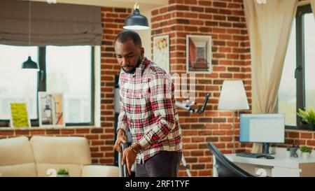 Personne masculine moderne faisant session de nettoyage de printemps à la maison, en utilisant un aspirateur pour nettoyer l'appartement. Jeune homme joyeux aspirant les planchers de salon en bois, en utilisant une solution de lavage. Banque D'Images