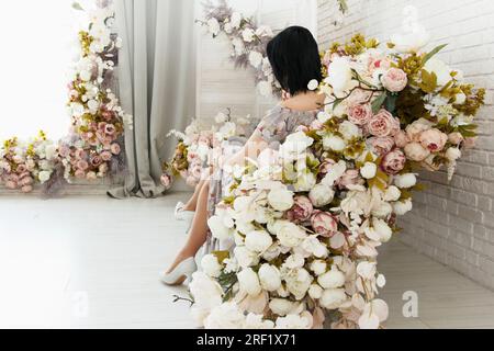 Fille élégante avec les cheveux foncés dans une robe et des chaussures se trouve à l'intérieur décoré belle avec des fleurs multicolores. Concentrez-vous sur l'épaule Banque D'Images