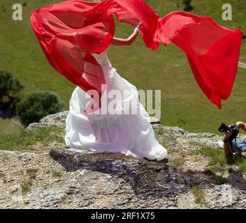 Mariée en robe blanche, tenant une écharpe rouge et posant sur le bord du canyon, photographe se trouve sur le sol au milieu de belles gorges et photographies mariée Banque D'Images