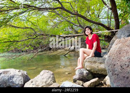 Jeune femme assise et se détendant sur de grands rochers dans la baie tranquille au large de l'océan hawaïen sous les arbres d'ombre Banque D'Images