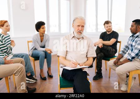 Portrait d'un thérapeute adulte senior confiant regardant la caméra tout en écrivant dans le rapport sur le presse-papiers. Patients multiculturels discutant dans Banque D'Images