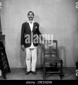 Vieux vintage noir et blanc des années 1900 photo de l'homme indien drôle portrait studio portant une chaise manteau debout Inde des années 1940 Banque D'Images