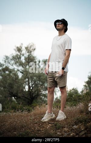 Un homme de 20 ans, vêtu d'un t-shirt blanc, d'un short beige, de lunettes de soleil et d'une casquette de marin noire, regardant sérieusement le ciel. Il est entouré par le Banque D'Images