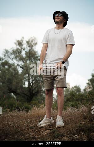 Un homme dans la vingtaine, riant ouvertement dans un t-shirt blanc, un short beige, des lunettes de soleil et une casquette de marin noire. Il est entouré par la beauté naturelle de Banque D'Images