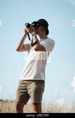 Portrait d'un homme d'une vingtaine d'années, habillé de façon décontractée d'un t-shirt blanc, d'un short beige, de lunettes de soleil et d'une casquette de marin noire, avec un camouflage classique Banque D'Images