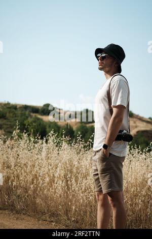 Portrait d'un homme d'une vingtaine d'années, habillé de façon décontractée d'un t-shirt blanc, d'un short beige, de lunettes de soleil et d'une casquette de marin noire, avec un camouflage classique Banque D'Images