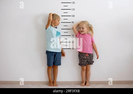 Petits enfants, garçon et fille blonds, mesurant la hauteur contre le mur dans la chambre, se comparant Banque D'Images