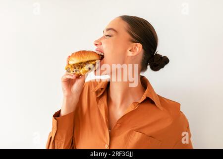 Satisfait jeune femme mordant savoureux Burger avec plaisir, appréciant le repas de triche sur fond de mur blanc Banque D'Images