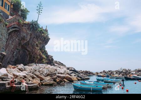 Cinque Terre, Italie - quai à Riomaggiore avec des bateaux et des touristes en maillots de bain. Ville balnéaire sur la Riviera italienne. Les gens sur une plage rocheuse sous le soleil Banque D'Images