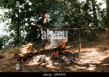 Voyage une bouilloire sur un feu brûlant sur la rivière et coucher de soleil backgroun. Cuisiner sur un feu de camp. Banque D'Images