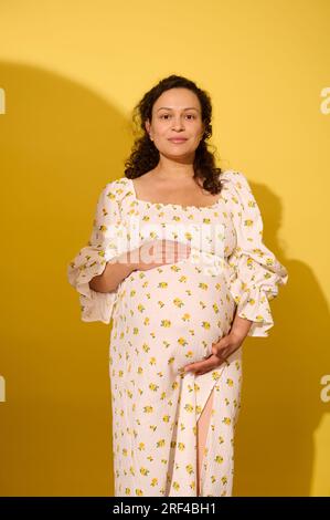 Charmante brune frisée, femme enceinte en robe blanche d'été, caressant son ventre au neuvième mois de grossesse heureuse et insouciante, souriant mignon lo Banque D'Images