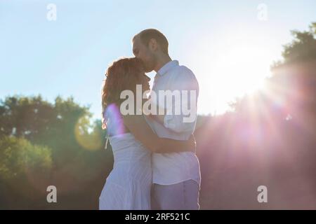 Photo créative de la Saint-Valentin de couple international, lumière du soleil, ciel, arbres sur fond, hommes caucasiens romantiques câlins, baisers front de latin Banque D'Images