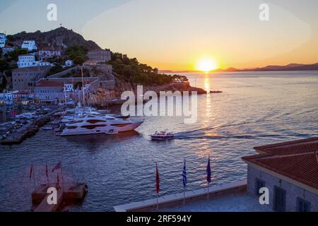 Baie d'Hydra dans un coucher de soleil fantastique. Petit bateau (bateau-taxi) de la Grèce continentale approchant du port. Grands yachts dans les parkings. Vacances idylliques Banque D'Images