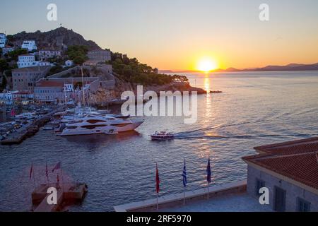 Baie d'Hydra dans un coucher de soleil fantastique. Petit bateau (bateau-taxi) de la Grèce continentale approchant du port. Grands yachts dans les parkings. Vacances idylliques Banque D'Images