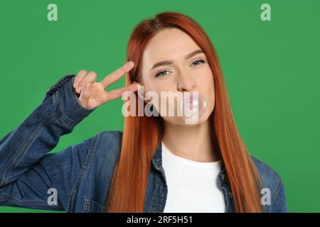 Belle femme soufflant du bubble gum et montrant le geste de paix sur fond vert Banque D'Images