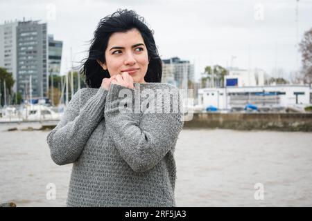 jeune belle femme latine touriste avec froid enroulé vers le haut, debout sur la jetée regardant sur le côté, avec l'eau de la rivière en arrière-plan, les gens et Banque D'Images