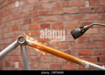 Utilisation d'un chalumeau pour enlever la peinture d'un cadre de vélo orange en cours de rénovation dans un atelier de vélo. Banque D'Images