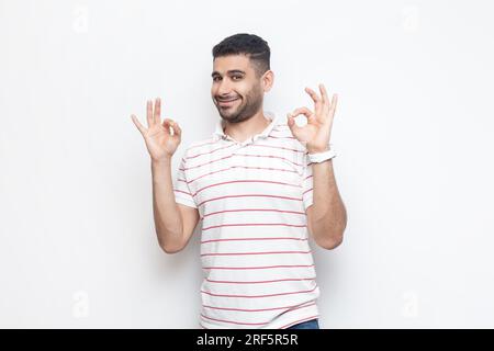 Tout va bien. Portrait d'un homme barbu souriant positif attrayant portant un t-shirt rayé debout montrant un geste correct, accepté. Studio intérieur tourné isolé sur fond gris. Banque D'Images